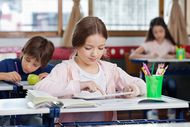 Student using her tablet at her desk. Two other students are in the background working at their desk. 